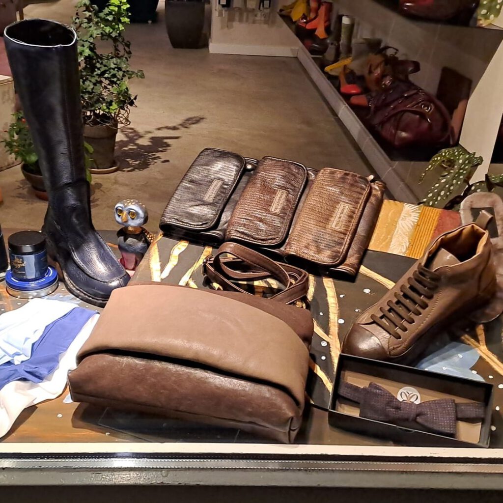 Ein dunkelblauer, durchgenähter Stiefel neben Burgol zur Pflege. Dazu ein passendes Set in Brauntönen, bestehend aus einem klassischen Schuh mit paralleler Schnürung und einer passenden weichen Clutch-Tasche, langen Geldbörsen und einer Fliege.