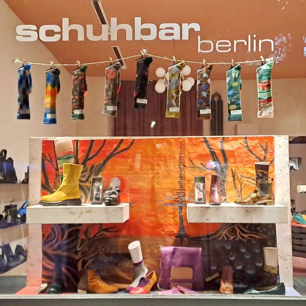 Schaufenstergestaltung mit handgemaltem Fantasy-Hintergrund, das schuhbar berlin logo auf der Scheibe, hängenden Kniestrümpfen sowie einer Auswahl an Stiefeletten, Tasche, Fliegen und Socken.