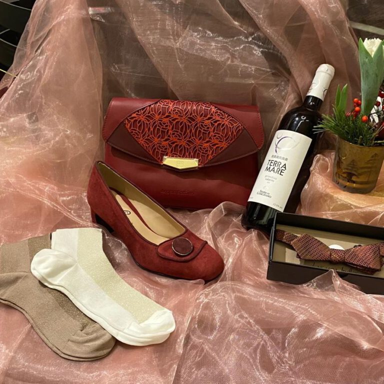 Ein roter Samtschuh, einer roten Ledertasche und einer roten Fliege auf zartrosa Organza. Ergänzt durch zwei Paar Socken in Weiß und Beige mit glitzernden Details sowie einer Flasche Wein neben einer Pflanze in einer eleganten Boutique-Präsentation.