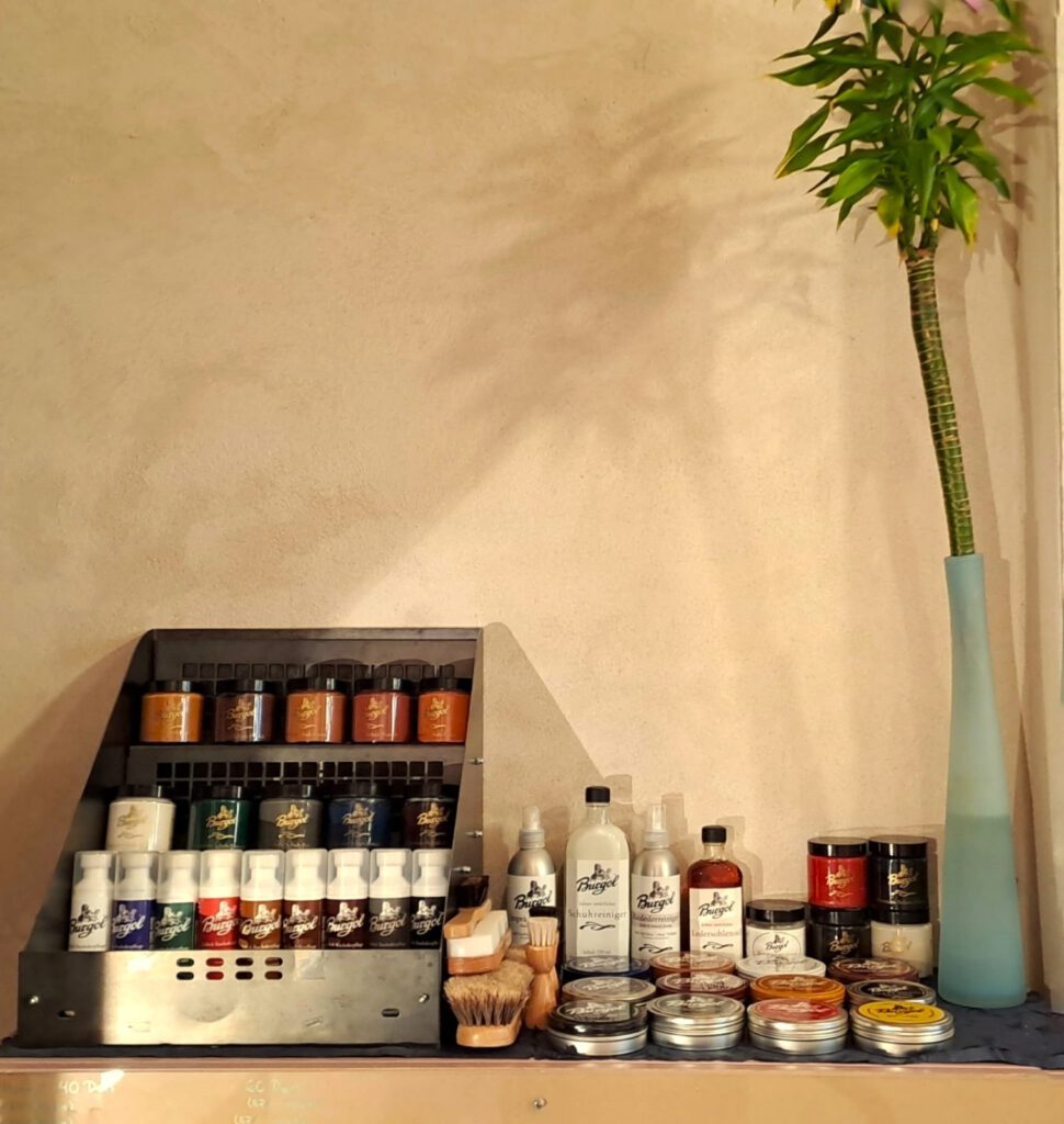 A selection of natural Burgol shoe care products including pomade, wax, cleaner, leather milk and sole oil arranged with a professional polishing brush made of animal hair.